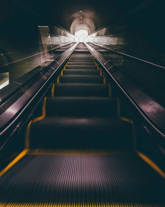 Escalator ascending into a bright tunnel, with yellow safety lines, in a dark, metallic environment.