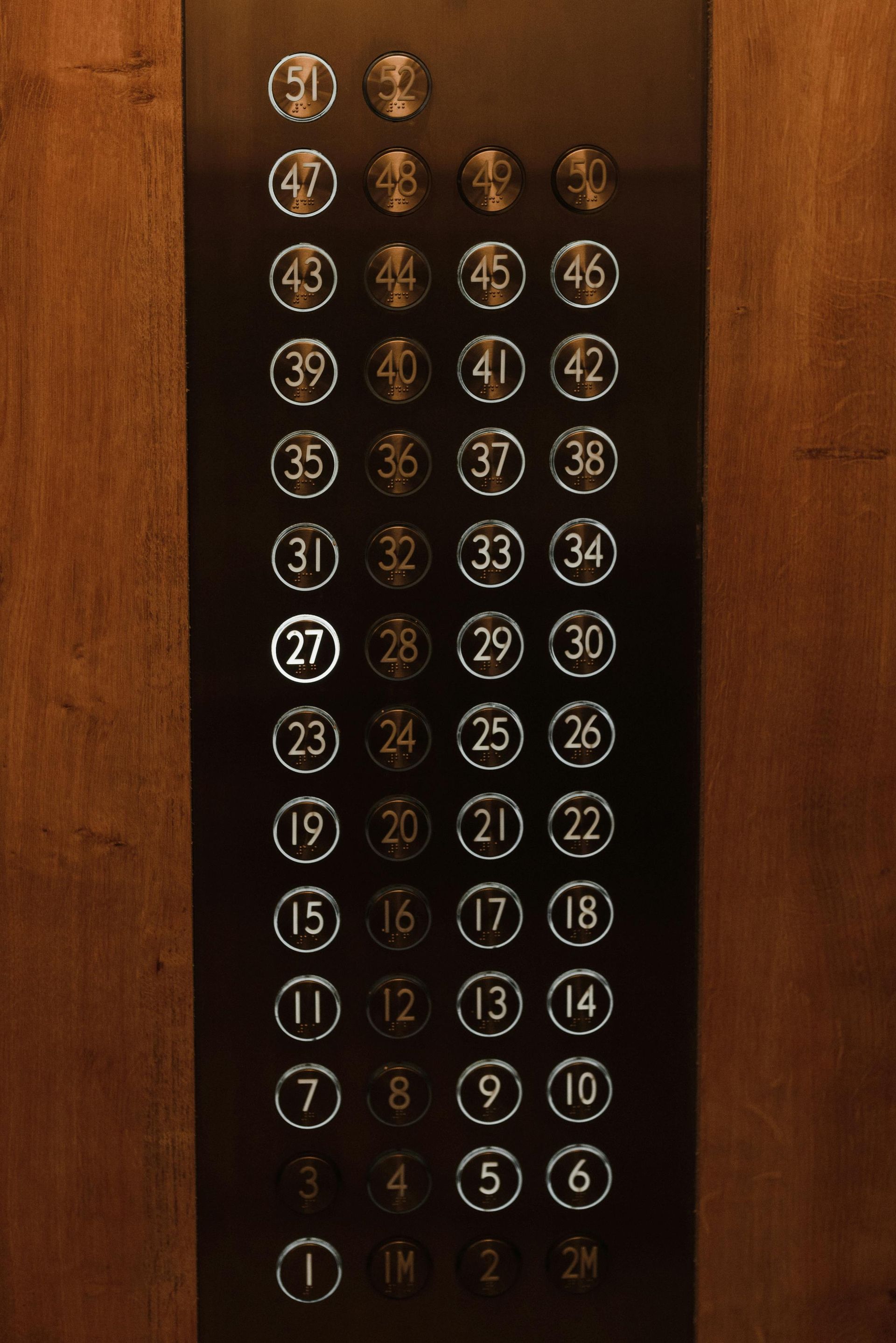 Elevator button panel with numbers 1-50, with button 27 illuminated. Wooden panel background.