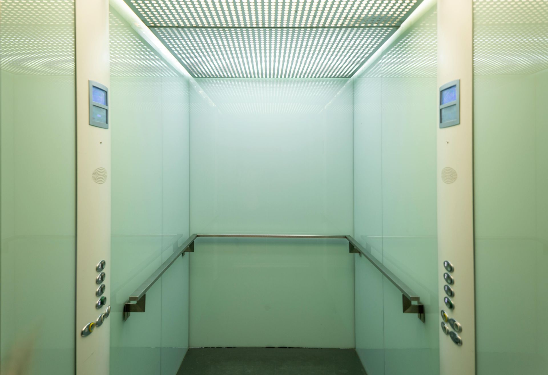 Empty elevator interior with handrails, buttons, and a grid ceiling, in light green and white tones.