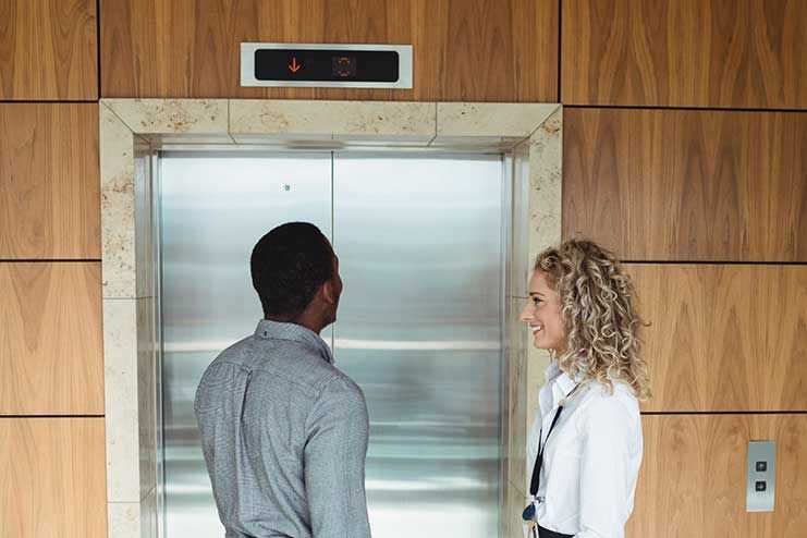 Two people waiting for an elevator with wooden paneling. The elevator's call button is lit.