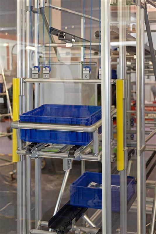 Vertical industrial lift with blue bins on conveyors within a metal frame. Yellow safety posts.