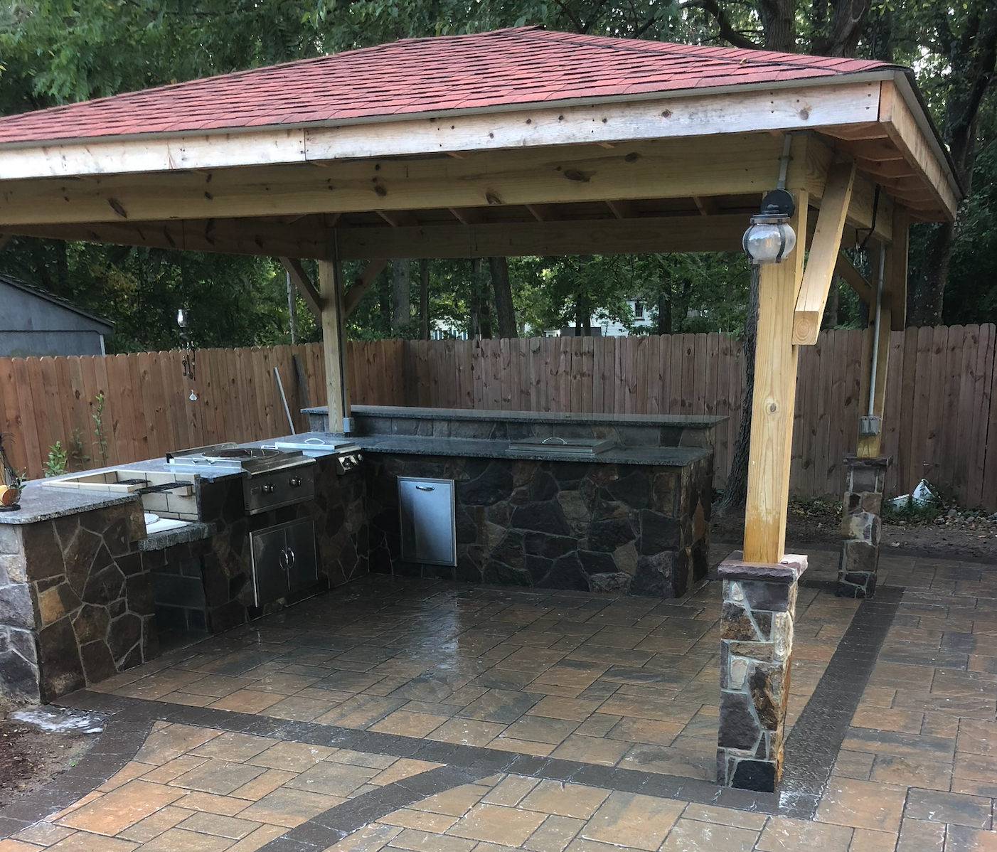 A wooden gazebo with a red roof is sitting on top of a brick patio. Outdoor kitchen inside gazebo