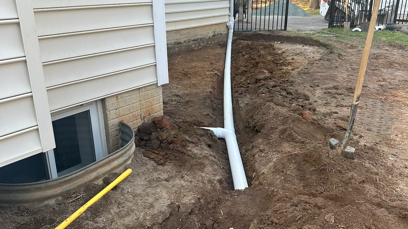 A white pipe is being installed in the dirt next to a house.