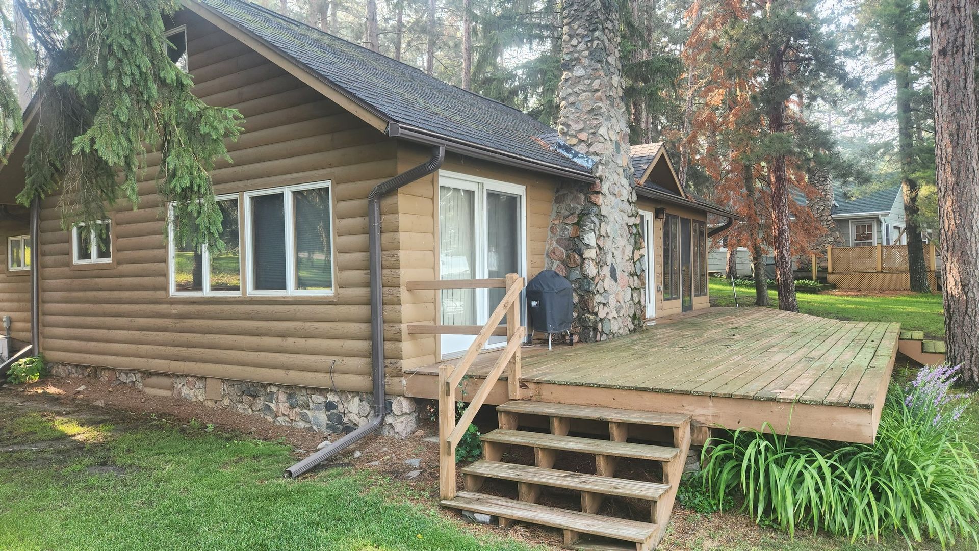 Log cabin with a wooden deck, surrounded by trees and greenery.