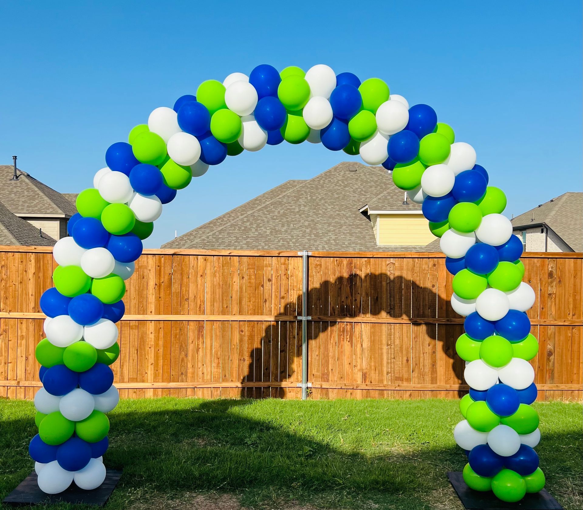 A blue green and white balloon arch in front of a wooden fence