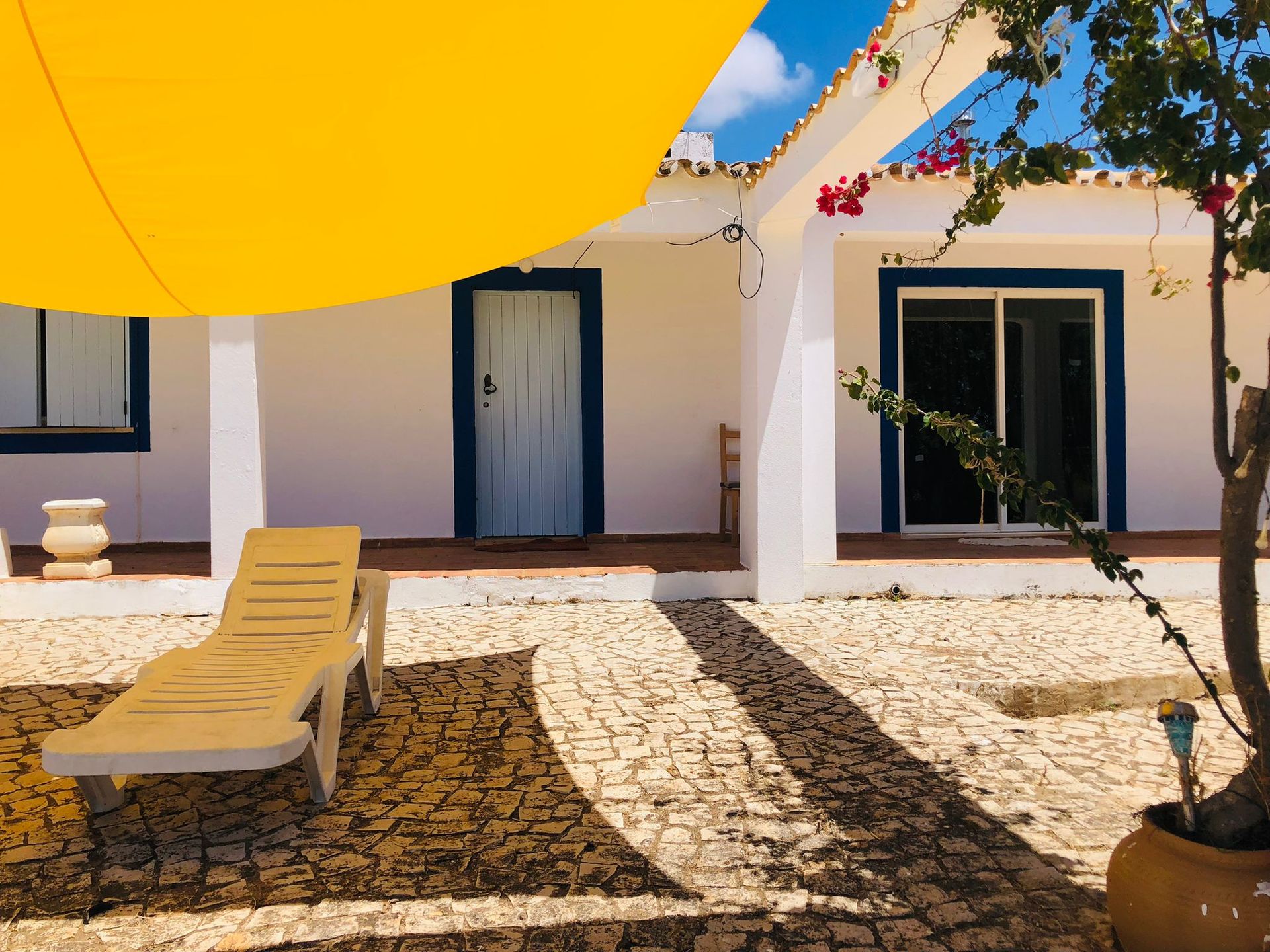 White house with blue trim, a yellow sunshade, and a beige chaise lounge on a stone patio.