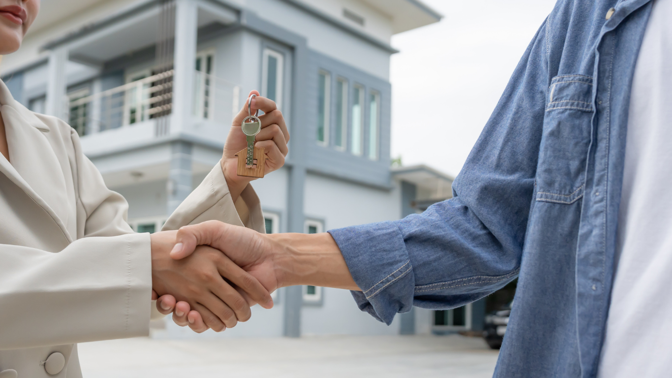 A professional in a beige suit shakes hands with another person, holding a house key in front of a modern home.