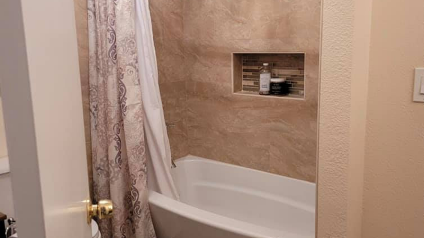A white bathtub inside a shower enclosure with beige tiled walls and a recessed shelf holding two bottles.
