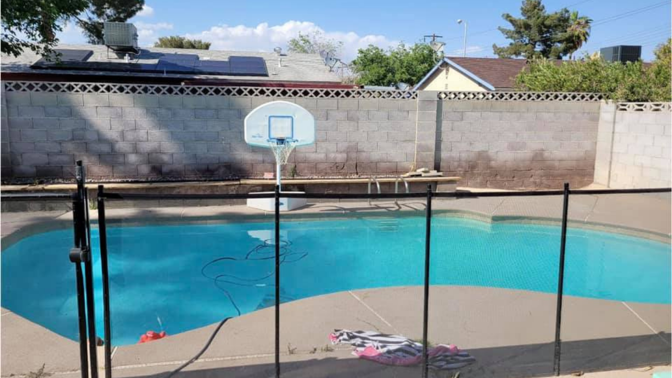 A backyard swimming pool with a turquoise interior, surrounded by a light-colored concrete deck and a black safety fence.