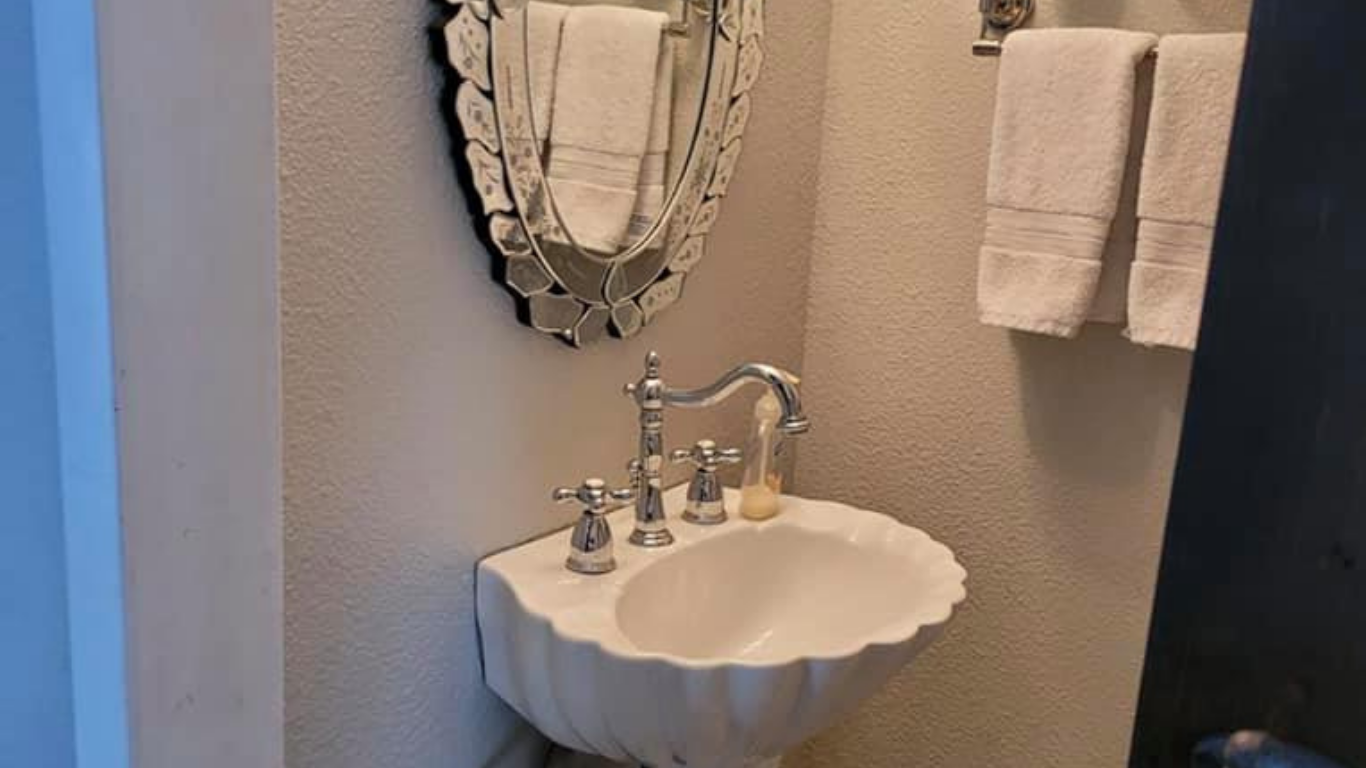 A white scallop-shaped wall-mounted sink with chrome hardware and an oval decorative mirror, flanked by hanging towels.