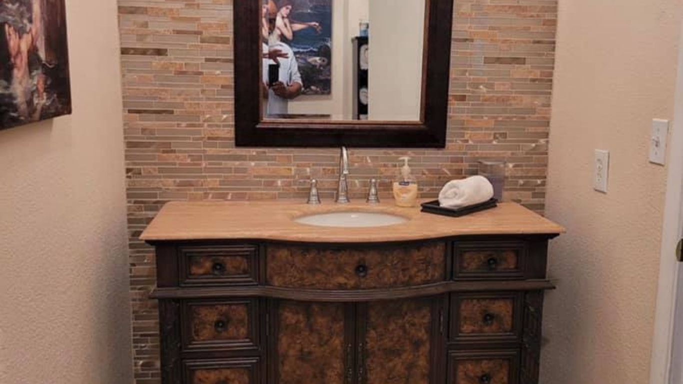 A bathroom vanity featuring a wooden cabinet, beige countertop, stone-tiled backsplash, and a framed mirror.