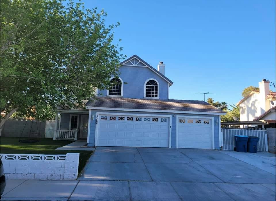 A two-story blue suburban house with a three-car garage, a concrete driveway, and a large tree in the front yard.