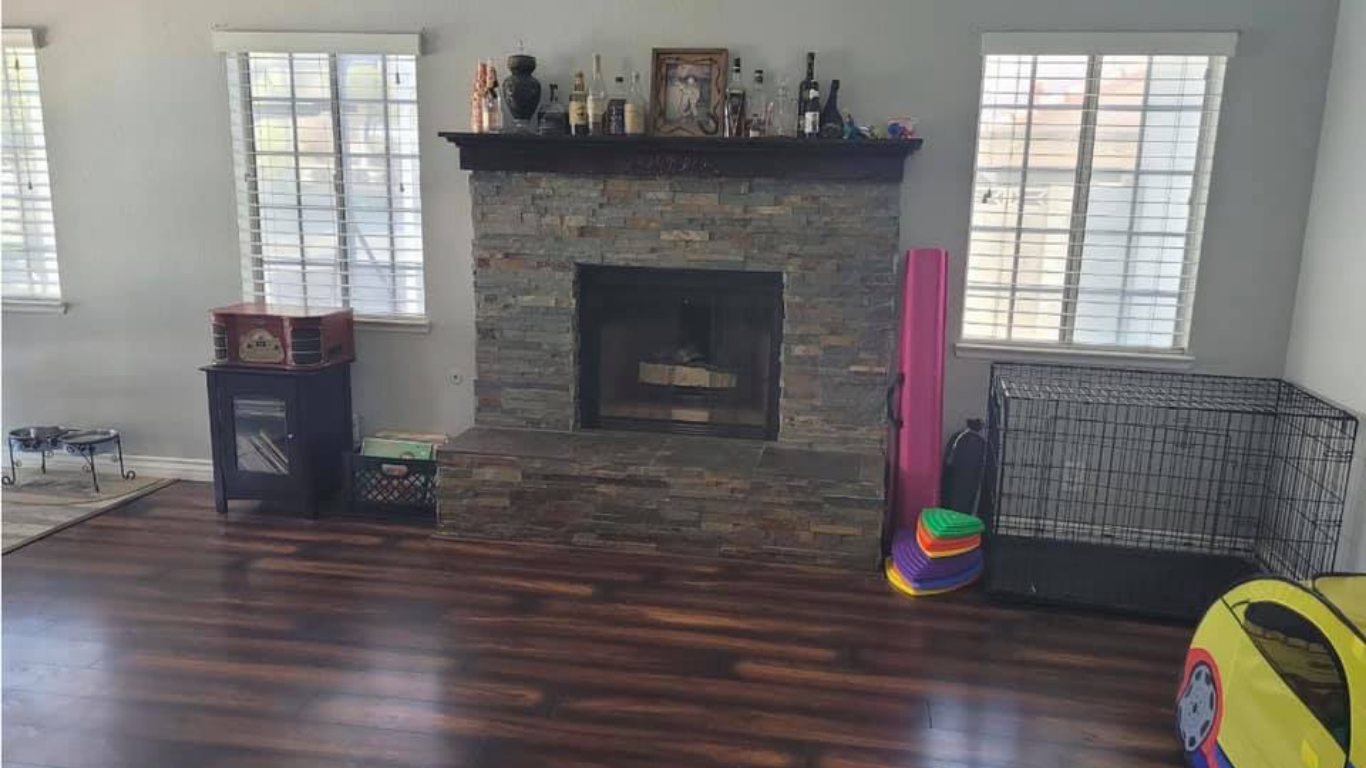 A stone fireplace sits between two windows in a room with dark wood flooring and various child-related items.