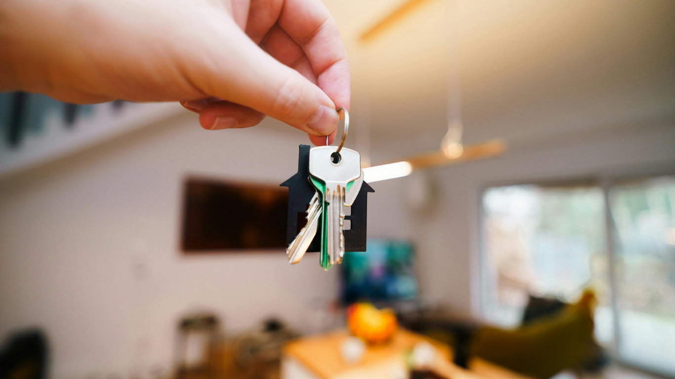 A close-up of a hand holding house keys with a black house-shaped keychain, with a blurred living room in the background.