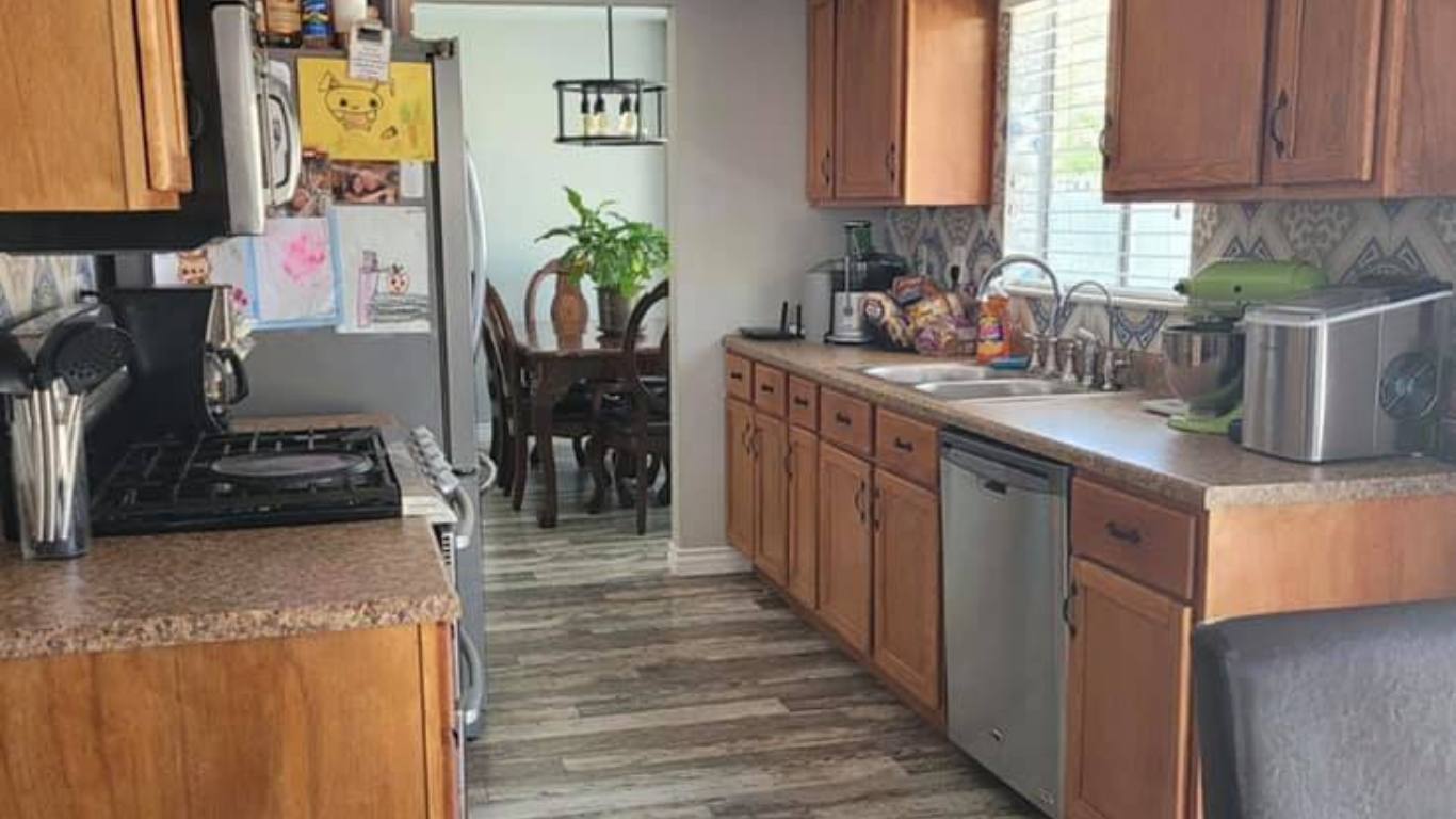 A kitchen with wooden cabinets, stainless steel appliances, and wood-look flooring, leading to a dining area with a plant.