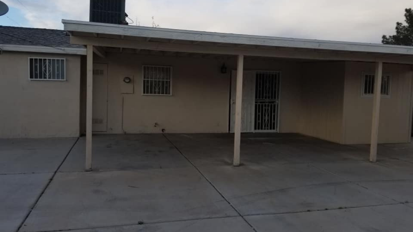 A beige house exterior featuring a covered concrete carport with support beams and security bars on the windows.