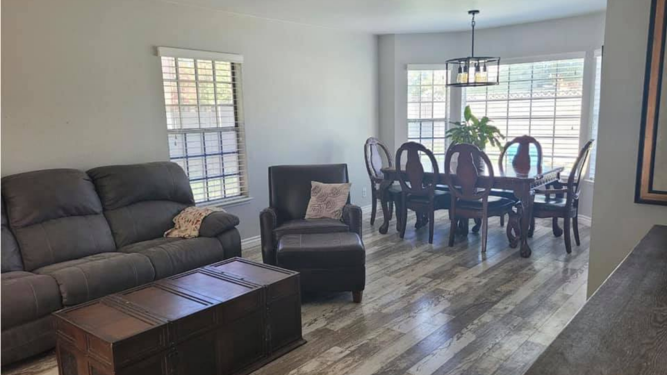 Living room with a gray sofa, a leather armchair with ottoman, a wooden chest table, and a dining set by a large window.