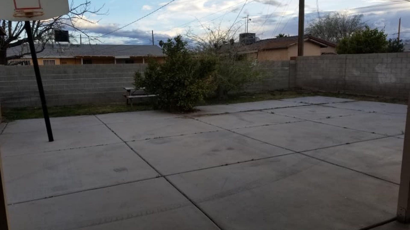 A concrete backyard patio featuring a basketball hoop on the left, a small tree against a back wall, and evening lighting.