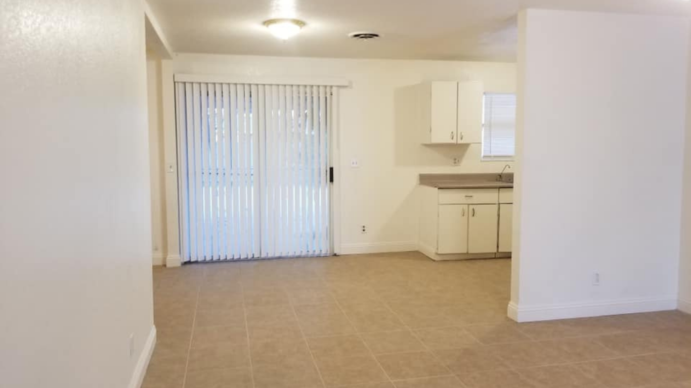 An interior view of a room with light-colored tile flooring, white walls, vertical blinds on a sliding door, and a kitchen.