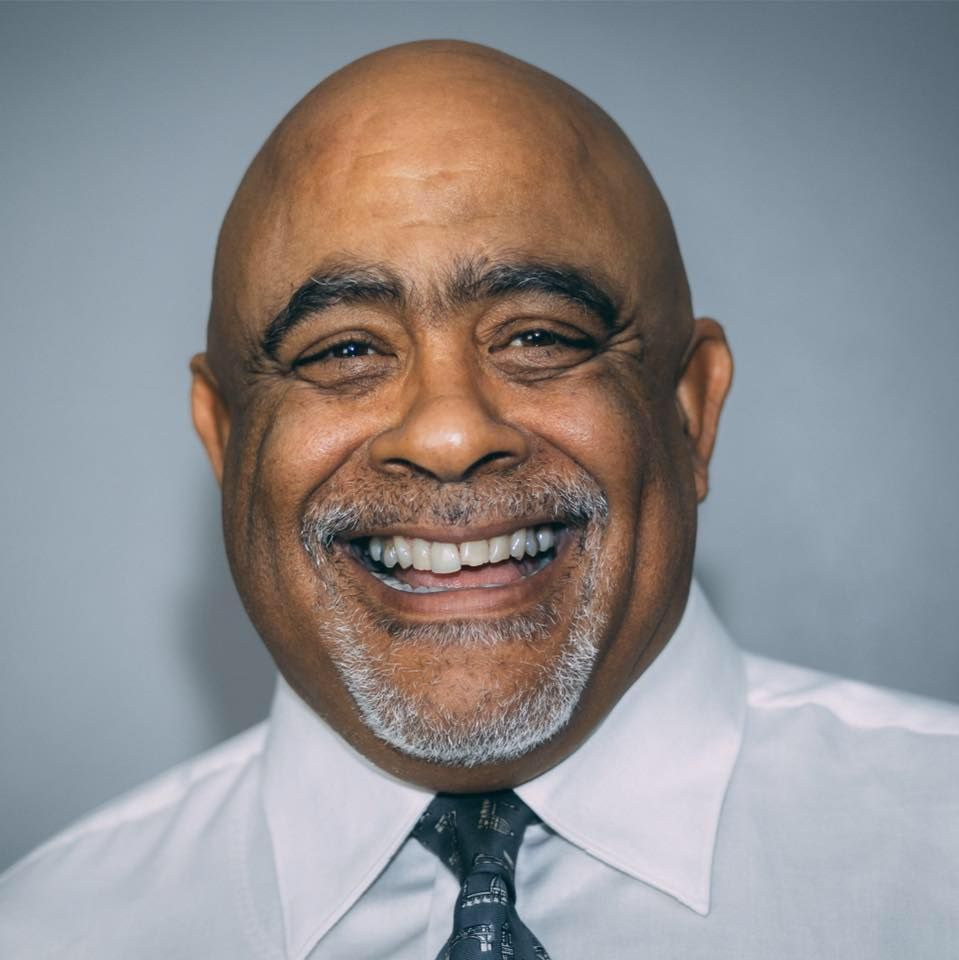 A bald individual wearing a white shirt and dark tie, smiling warmly against a plain gray background.