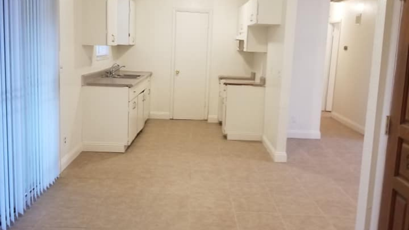 A brightly lit kitchen with white cabinets, a sink, and beige flooring, viewed from an adjacent open living space.