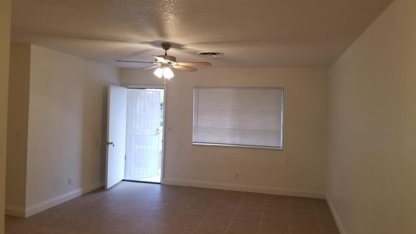 Empty, neutral-toned room with beige tile flooring, a ceiling fan, a window with closed blinds, and an open white door.