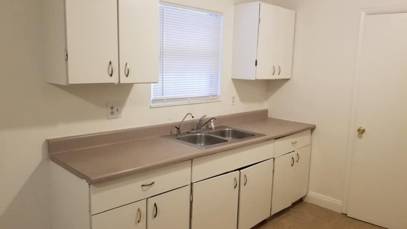 A kitchen area featuring a tan countertop, double-basin sink, and white cabinets against plain walls with a window.