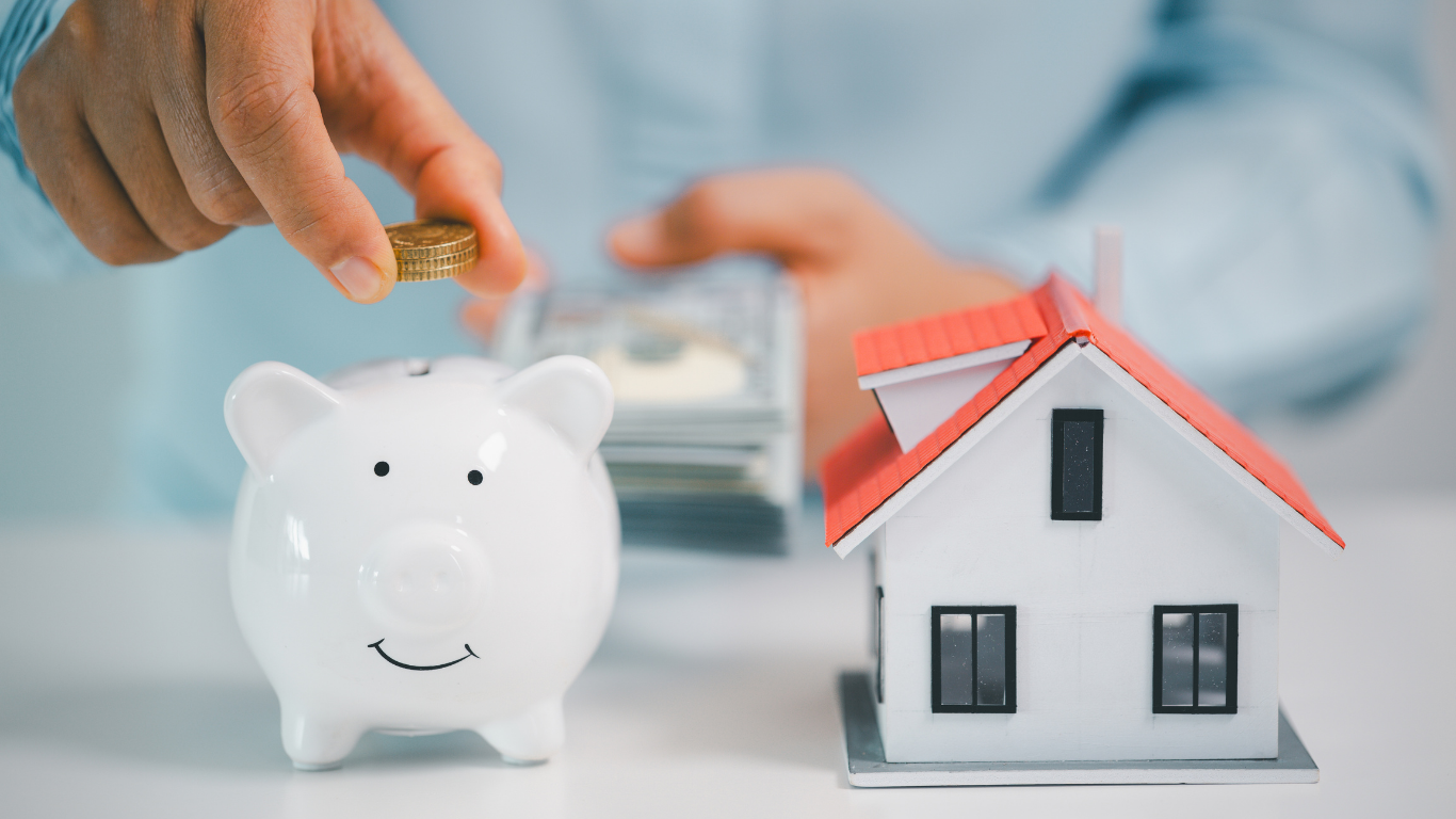 A hand places a coin into a white piggy bank next to a small model house and a stack of cash.