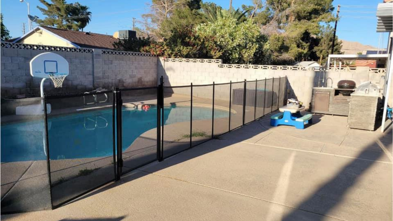 A black mesh safety fence surrounds an outdoor swimming pool on a concrete patio next to a basketball hoop.