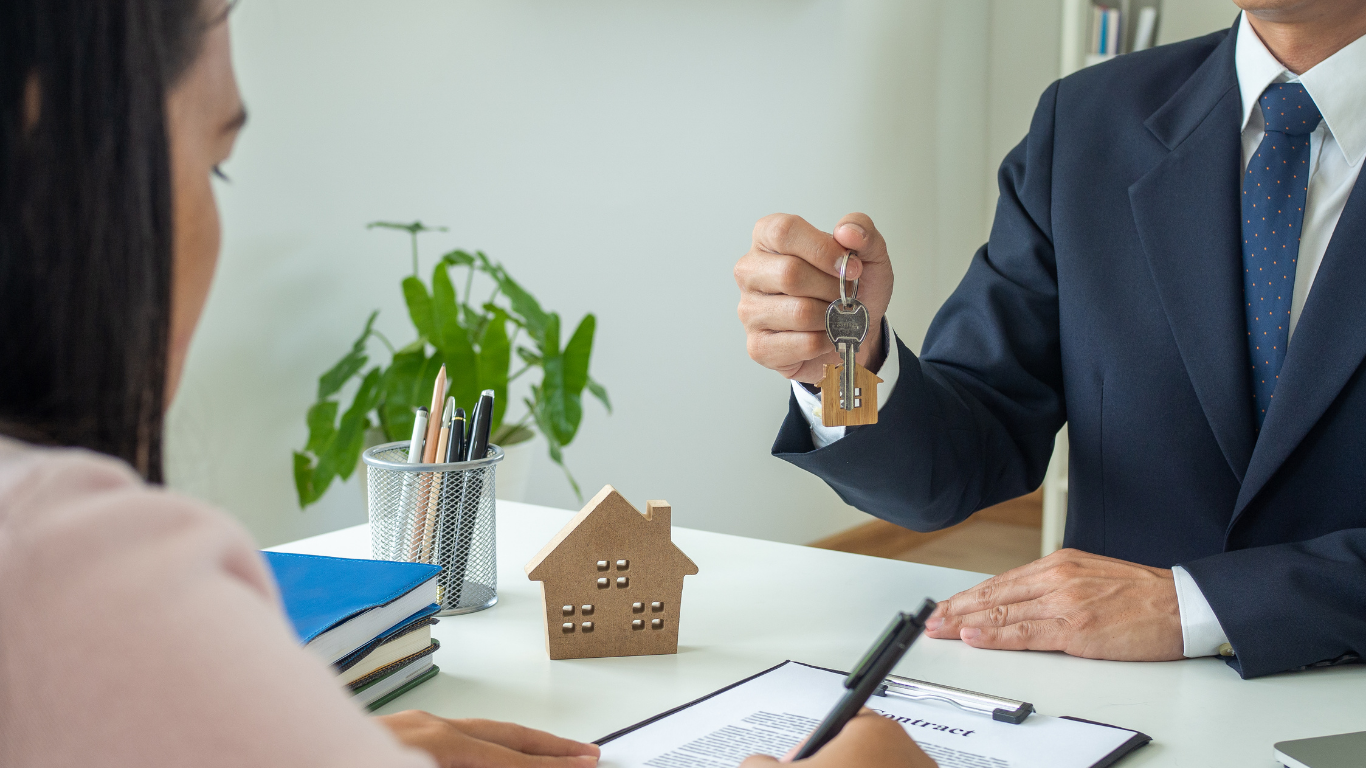 A professional in a suit hands keys to a client over a desk with a small wooden house model and contract.