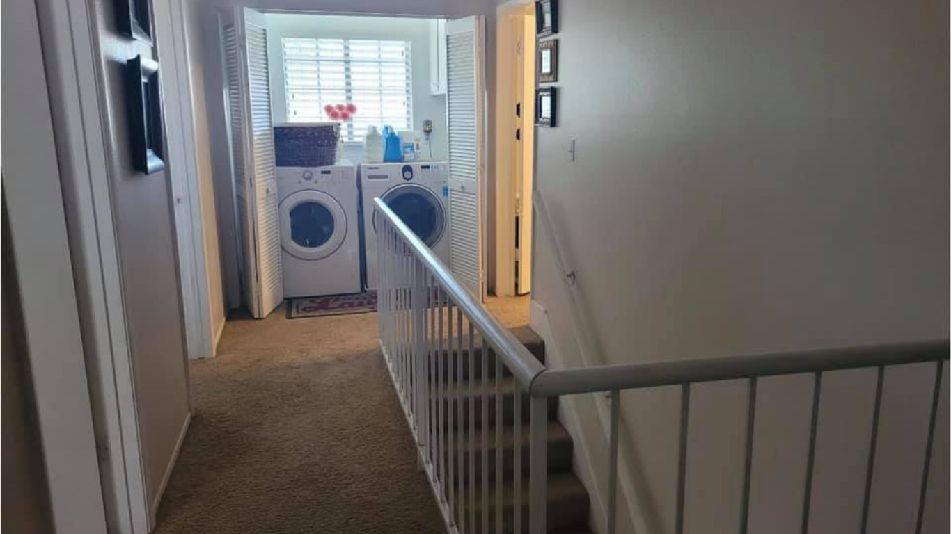 An indoor laundry area with a stacked washer and dryer at the end of a hallway, next to a staircase with a white railing.