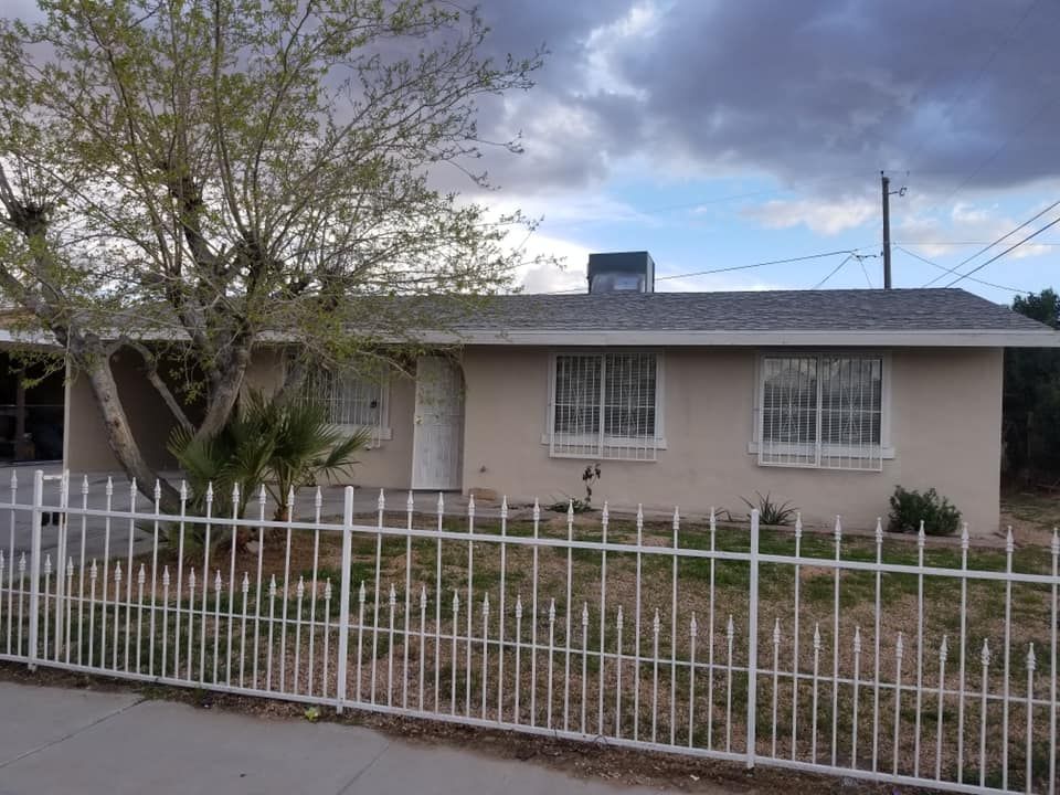 A single-story, beige stucco house with a gray roof and white metal security bars on the windows, behind a white fence.