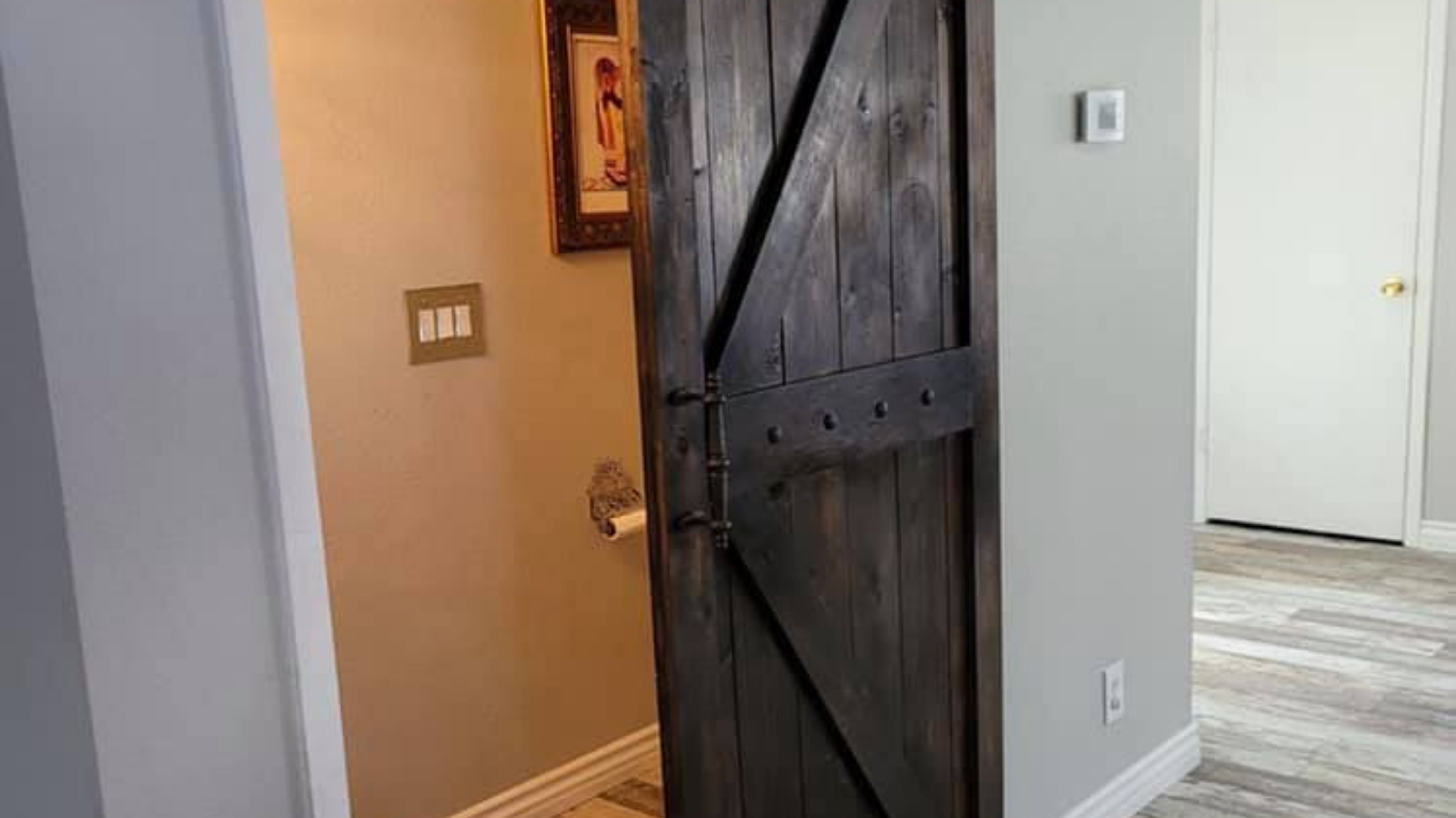 A dark, rustic wooden barn-style door stands partially open in a bright home interior with light walls and wood floors.