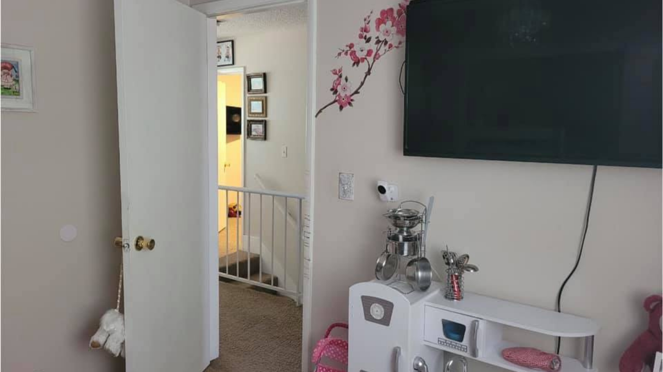 A light-colored room features a mounted television, a floral wall decal, a small toy kitchen, and an open doorway.