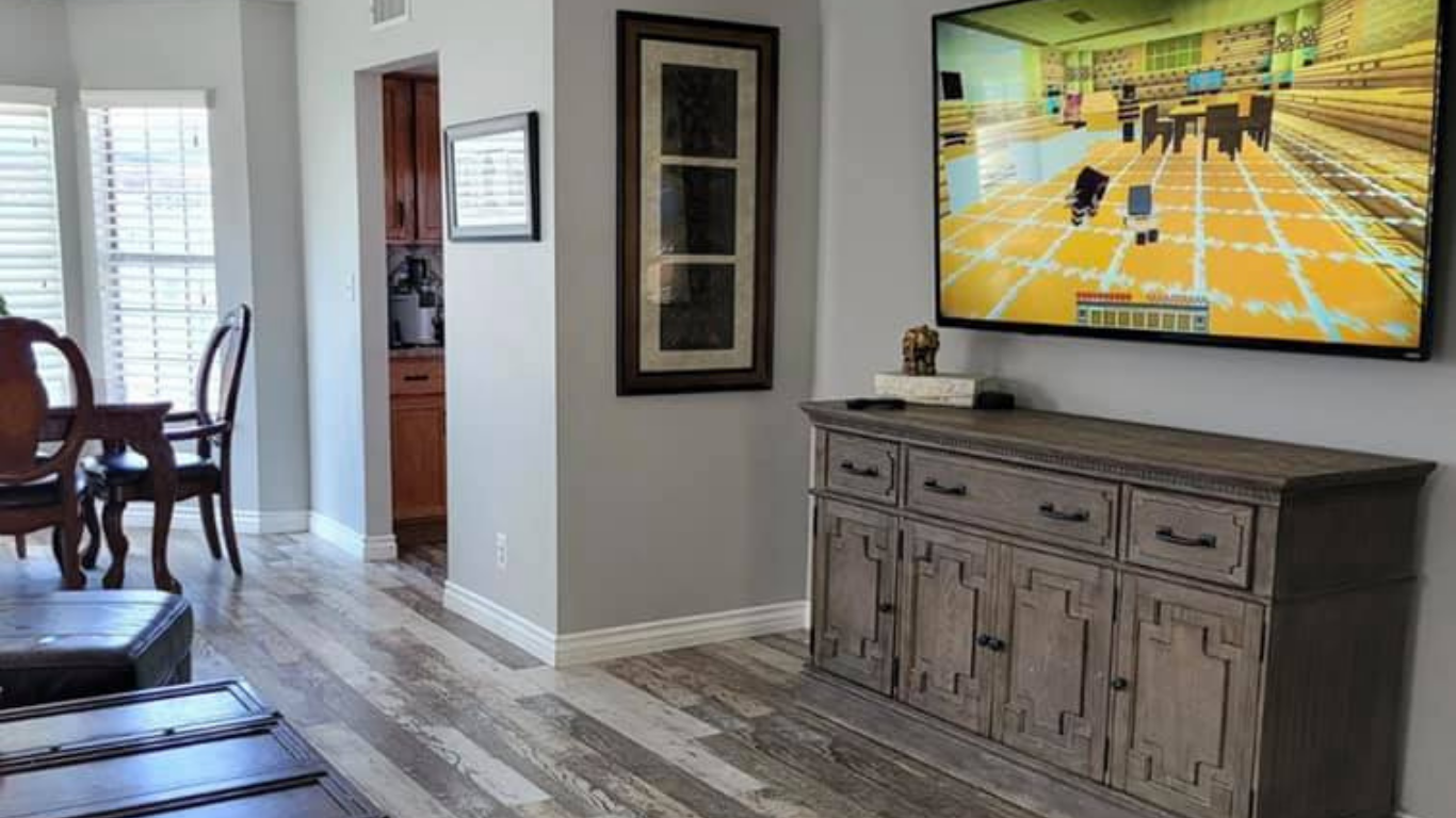 A living room featuring a TV showing a Minecraft game, a wooden sideboard, and a nearby dining area with chairs.