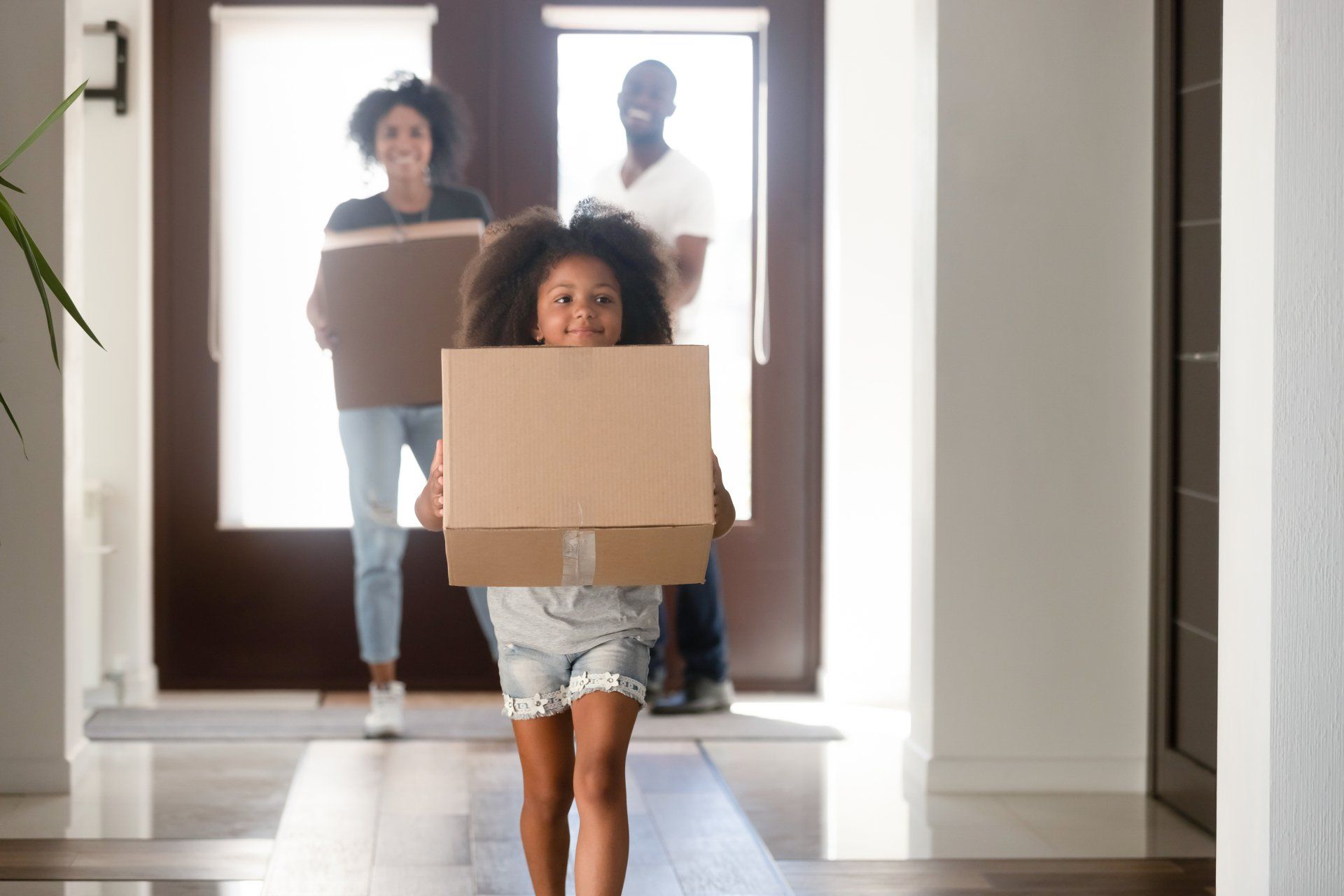 Family moving into a new home, carrying boxes. Child smiling in front, parents behind. Interior hallway.