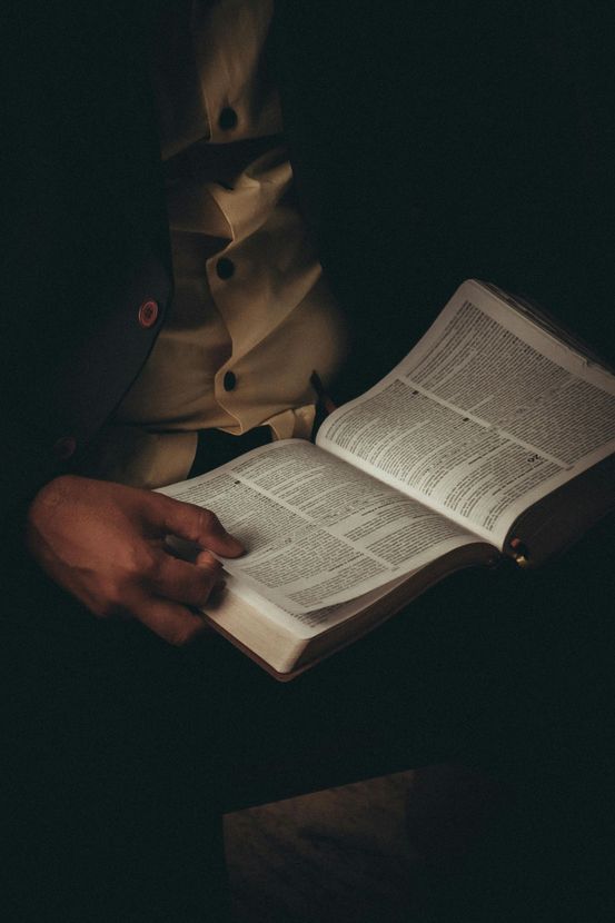 A person in a dark suit and light vest holds an open book, reading the printed text in low light.