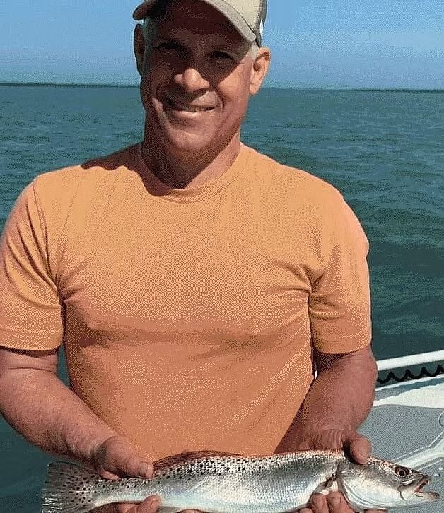 Man smiling while holding a spotted fish on a boat, water and sky in background.
