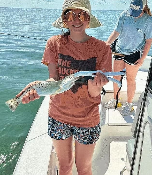 Woman on a boat smiling, holding a fish with pliers. Wearing hat, t-shirt, shorts. Another person stands behind.