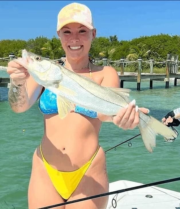 Woman in bikini holding a fish on a boat, smiling at camera. Beach in the background.