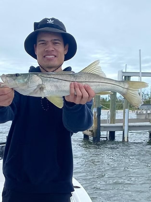 Man smiles, holding a silvery Snook fish, by a dock. He wears a black hat and a blue sweatshirt.