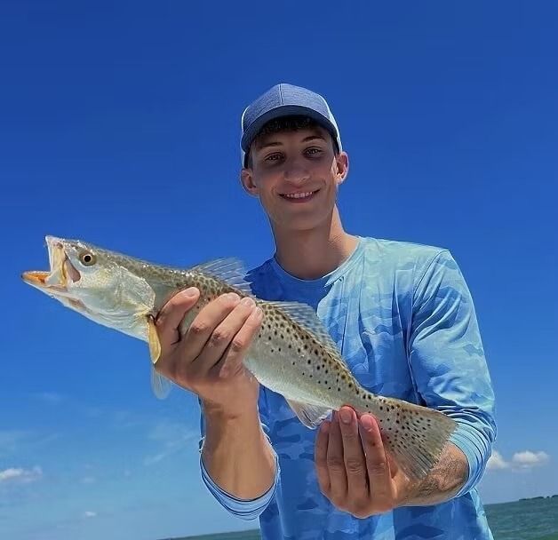 Young man in blue holds speckled trout against a bright blue sky, smiling.