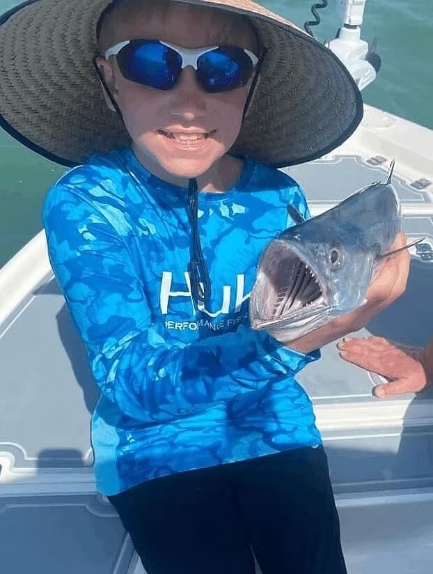 Boy on a boat smiles, holding up a fish with wide open mouth; wearing sunglasses and a sun hat.