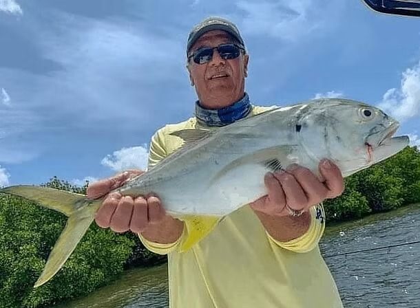 Man holding a large, silver fish with yellow fins. Blue sky and water background.