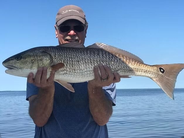Man holding large redfish in front of a blue sea, wearing sunglasses and a hat.