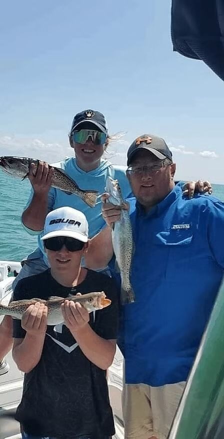 Three people on a boat, smiling, holding up freshly caught speckled trout, with blue sky and water in the background.