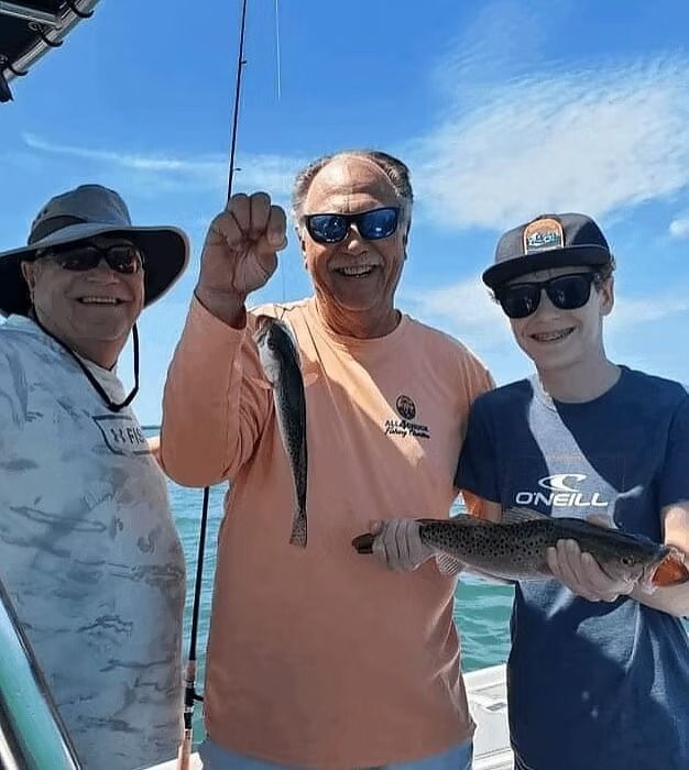 Three people on a boat holding up fish they caught, smiling. Ocean and blue sky in background.