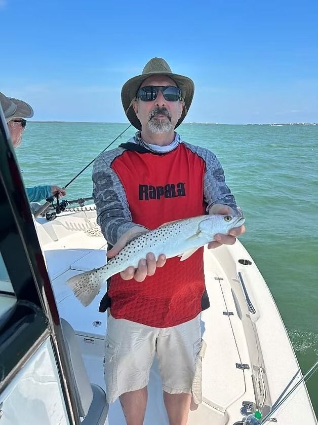 Man on a boat holding a speckled trout, wearing hat and sunglasses, under a blue sky.