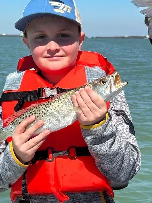 Boy in life jacket holds up spotted fish on a boat, smiling.
