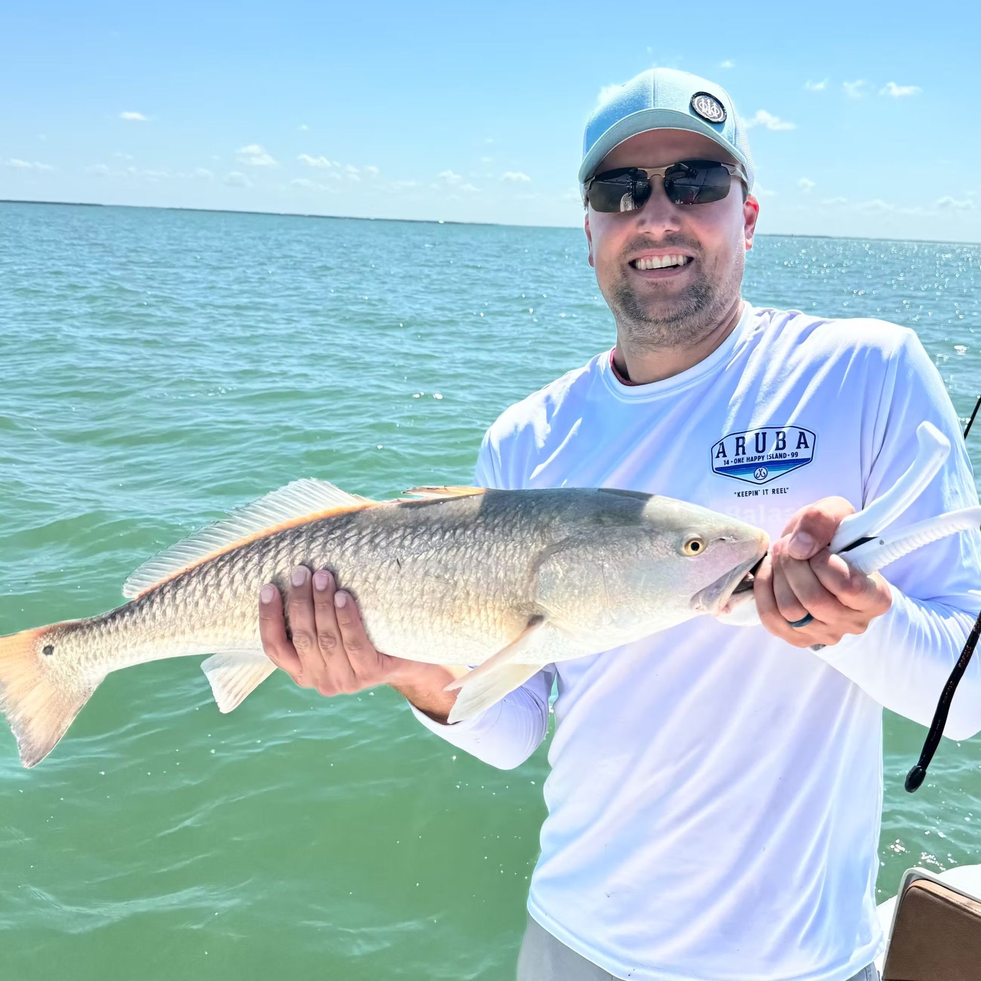 Man smiles, holding a large redfish on a boat, near the ocean on a sunny day.